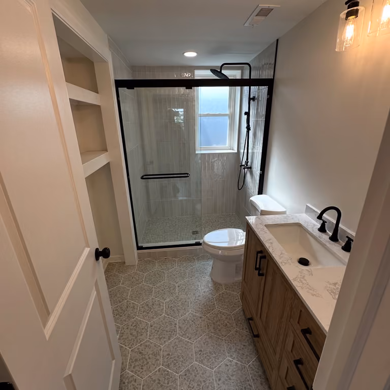Narrow remodeled bathroom with dark framed glass shower and long light-colored vanity.
