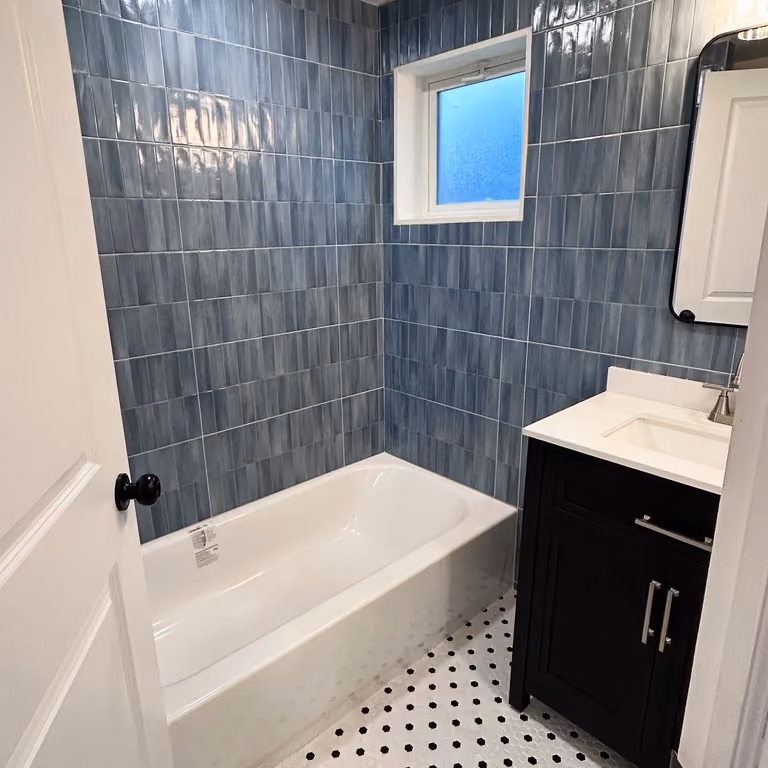 Bathroom with blue-gray tile, white bathtub, black hardware and light window.