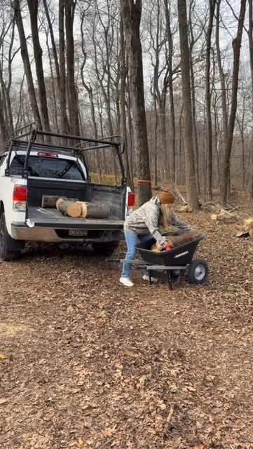 A person loading cut logs beside a white pickup in winter woodland.